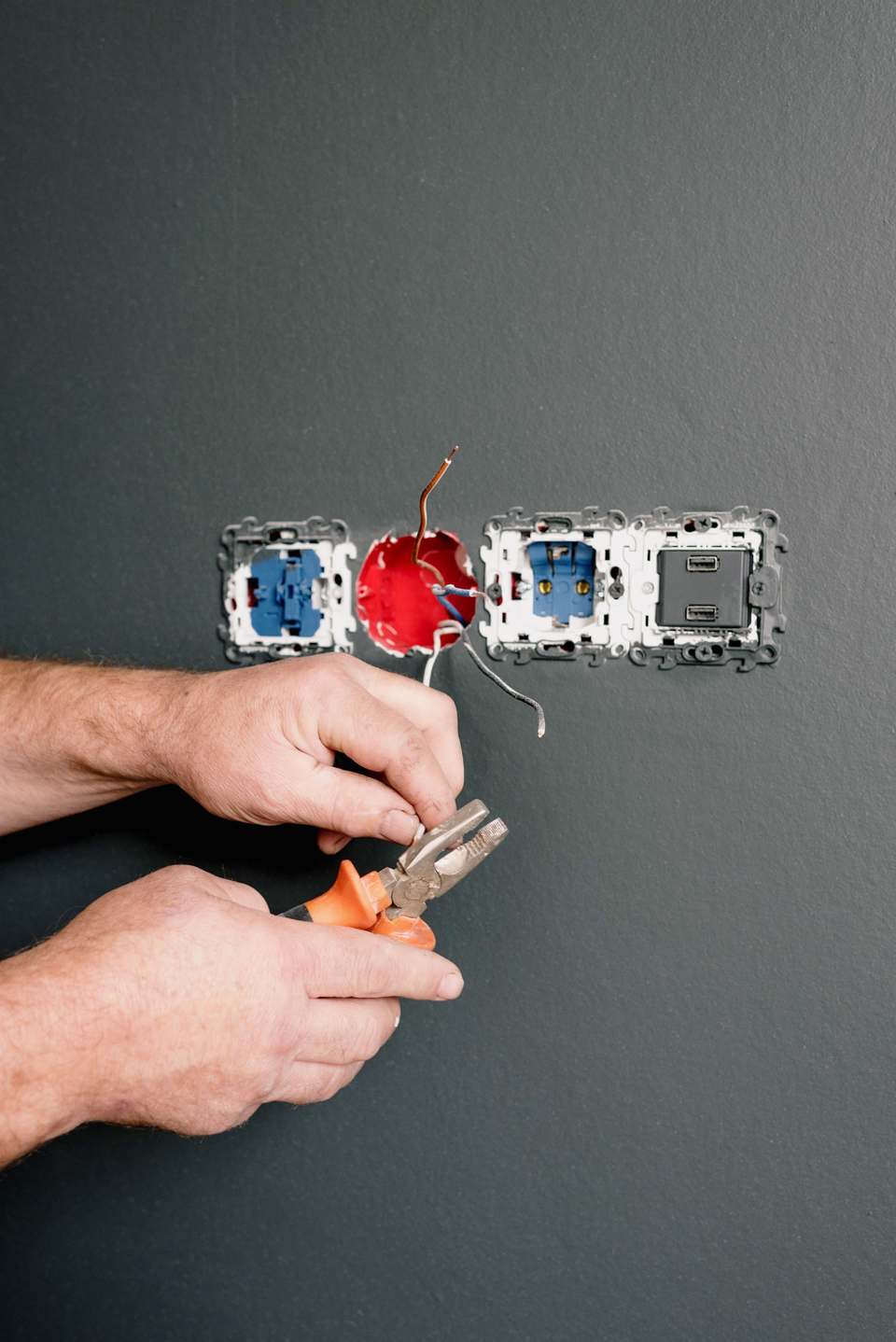 Electrician lucrând cu cleștele la firele unei prize, într-o cameră cu pereți gri închis
